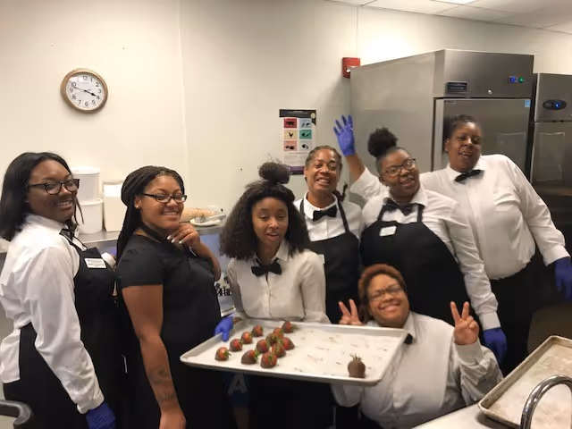 A group of six smiling kitchen staff members wearing white shirts, black aprons, and gloves pose together in a commercial kitchen. One person is holding a tray of chocolate-covered strawberries, and another is making a peace sign.
