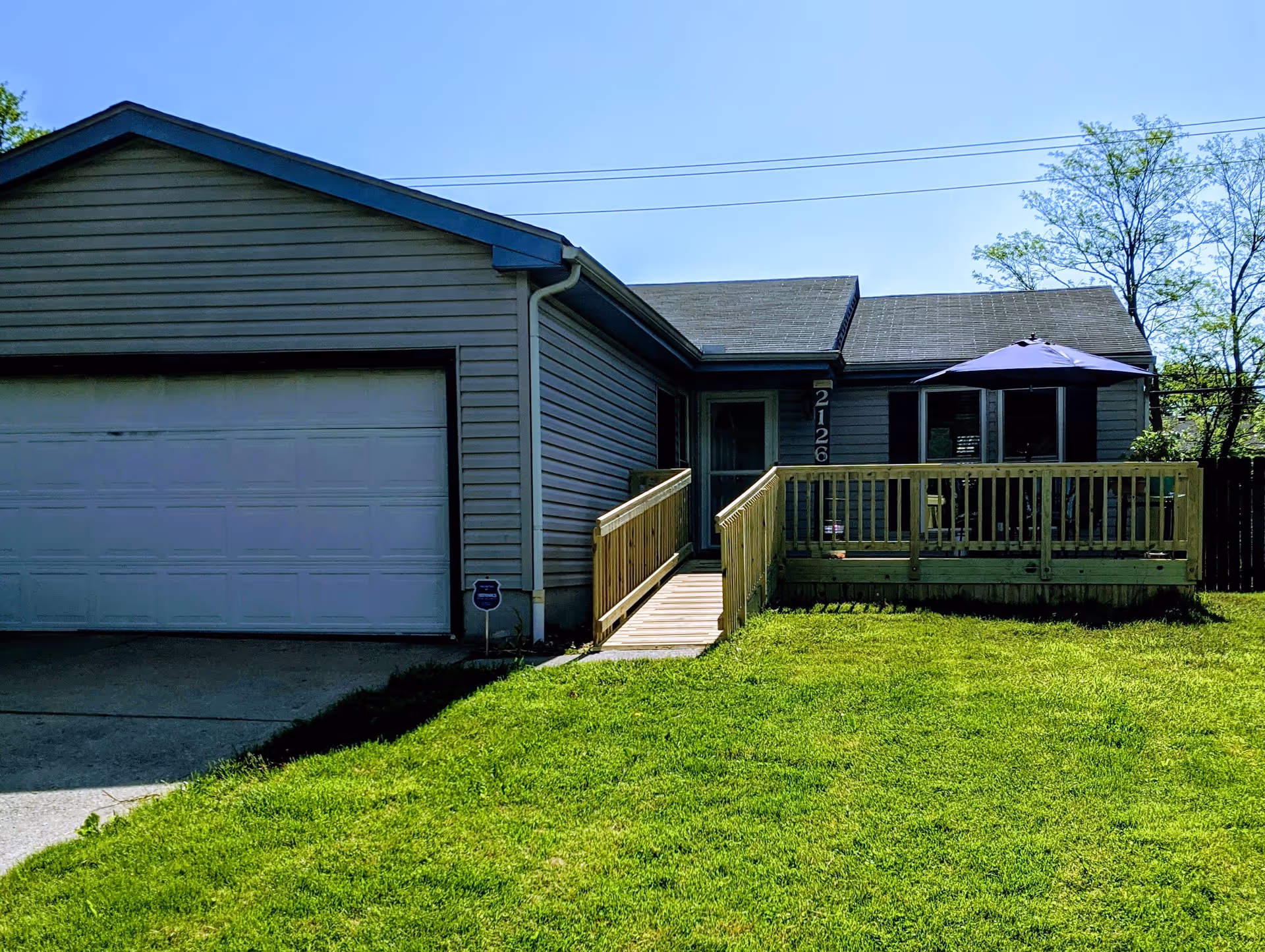 Exterior view of a single-story house with gray siding, a white garage door, a wooden ramp leading to the front door, and a wooden deck with a purple umbrella. The house number 2126 is visible near the entrance. The lawn in front is green and well-maintained, with trees in the background under a clear blue sky.