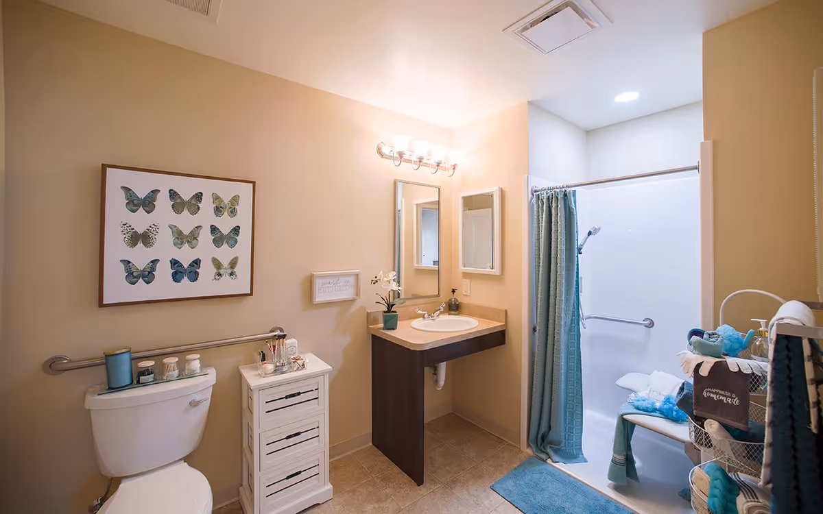 A well-lit bathroom with beige walls featuring a toilet with a grab bar, a small white cabinet with toiletries, a sink with a mirror above it, and a walk-in shower with a blue curtain and a bench inside. The bathroom is decorated with a framed butterfly artwork and a small sign on the wall.