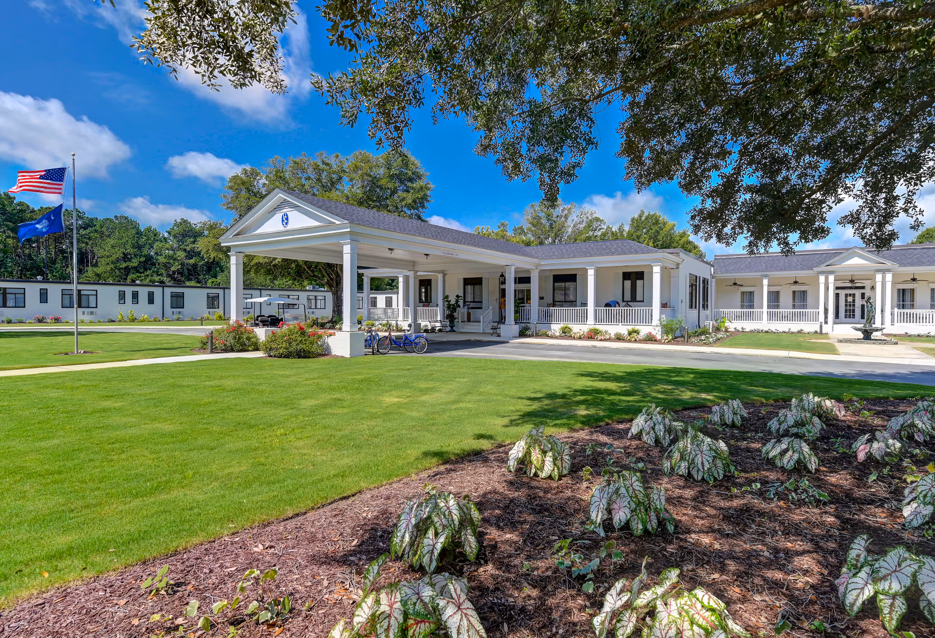 Exterior view of The Florence Presbyterian Community building with a covered entrance, well-maintained green lawn, landscaped garden with plants, and two flagpoles displaying the American flag and another flag under a bright blue sky with some clouds.