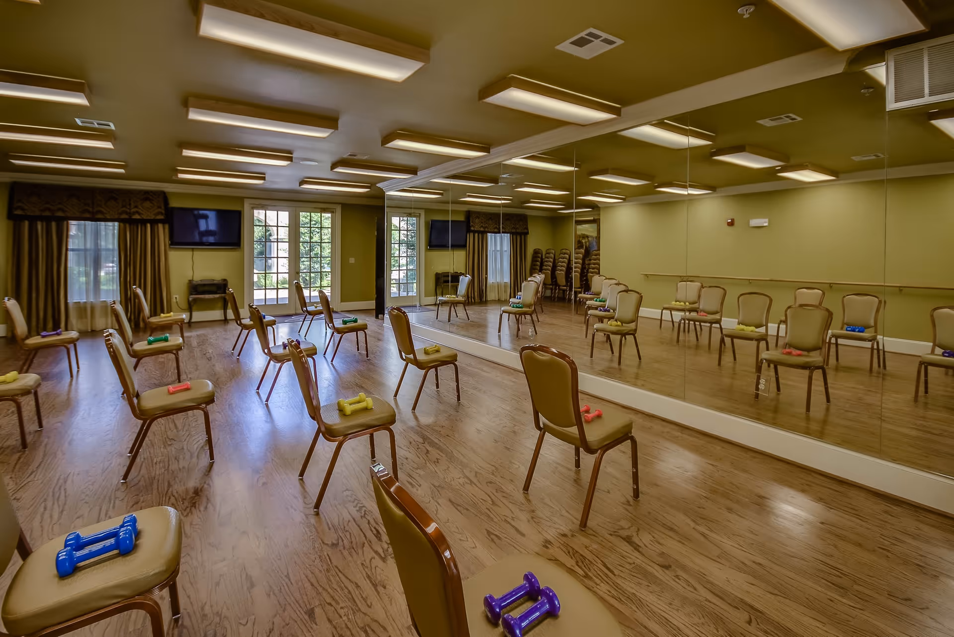 A spacious exercise room with wooden floors and green walls, featuring a large wall mirror. Several chairs are arranged in rows, each with a pair of colorful dumbbells placed on the seat. The room has large windows and glass doors letting in natural light, with two wall-mounted televisions and stacked chairs in the corner.