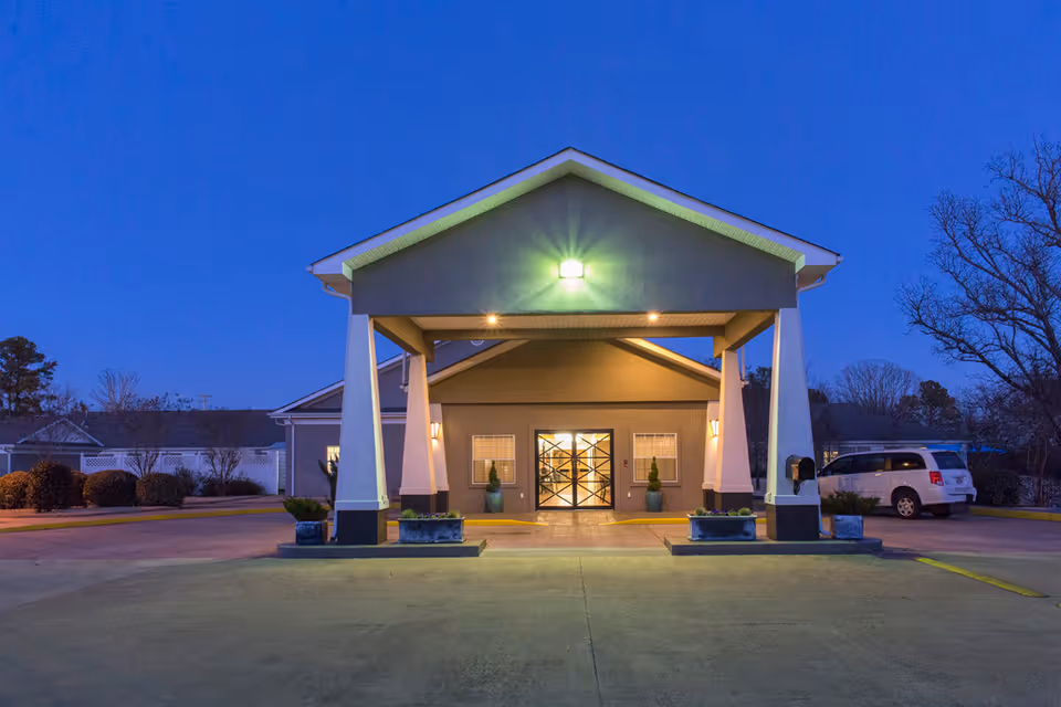 Front exterior view of Quapaw Nursing & Rehab Center at dusk with a covered entrance, illuminated lights, potted plants, and a parked white vehicle to the right.