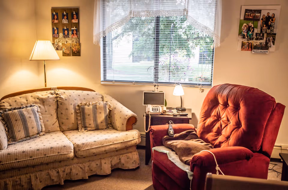 A cozy living room area with a beige patterned sofa with matching cushions on the left and a plush red recliner chair on the right. Between them is a small dark wooden side table holding a telephone, a small lamp, and some other items. Behind the furniture is a window with blinds and a lace valance, showing greenery outside. On the walls are photo collages and a floor lamp beside the sofa.