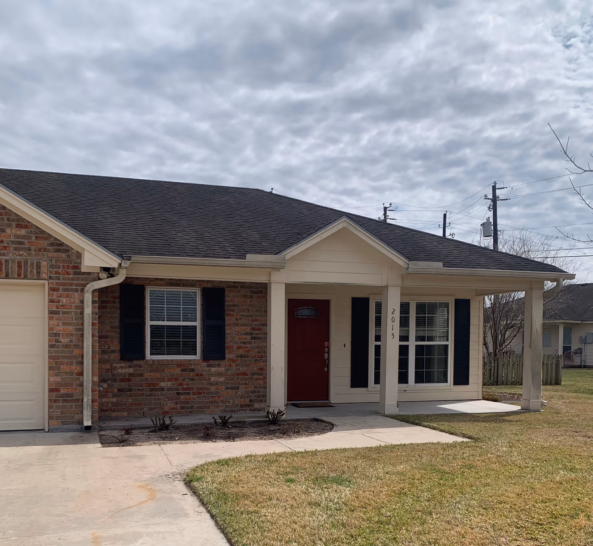Exterior view of a single-story brick and siding house with a dark shingled roof, a red front door, two windows with black shutters, a small covered porch supported by two columns, and a driveway leading up to the house. The house number 2015 is visible on one of the porch columns. The sky is cloudy and there is grass in the front yard.