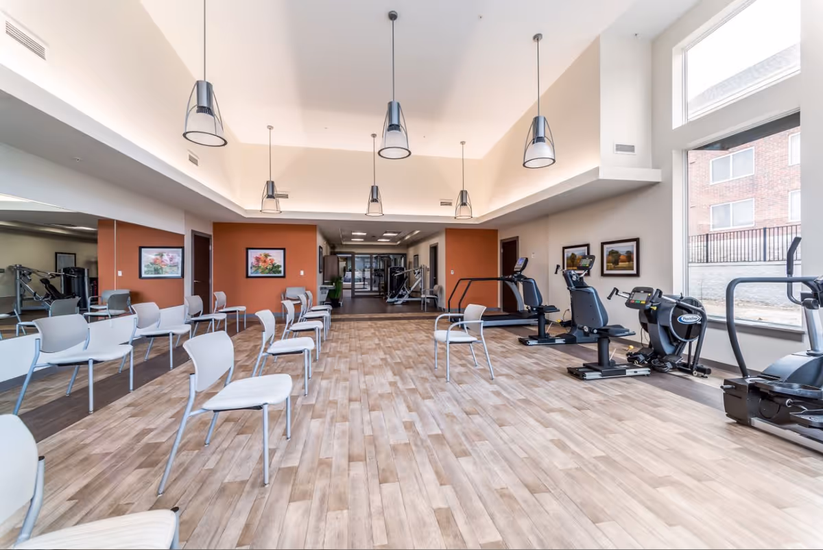 Spacious senior living fitness/activity room with rows of chairs, exercise machines, pendant lights, and large windows.