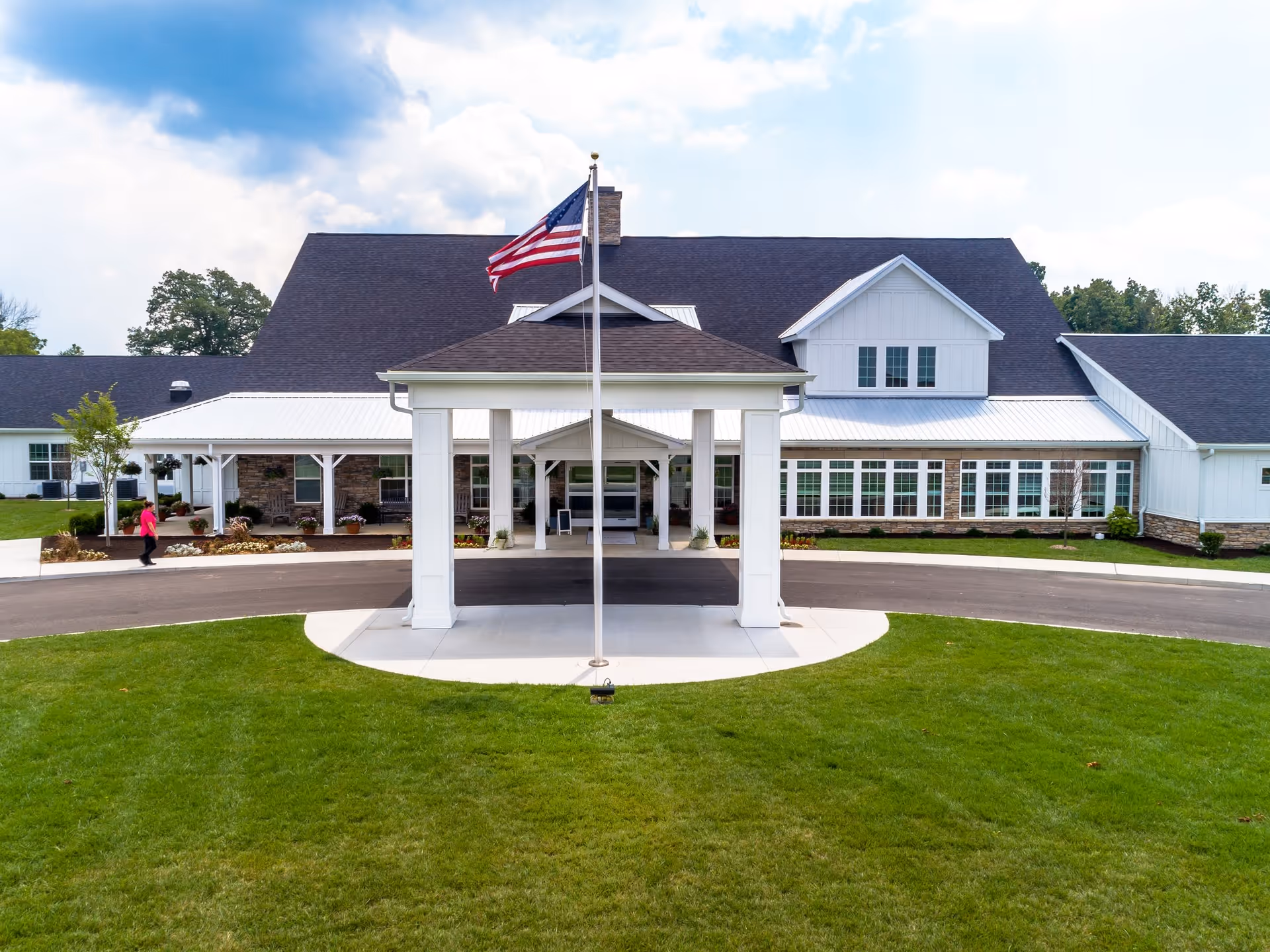 Front exterior view of a large senior living facility building with white siding and a dark roof. There is a covered entrance with white pillars and an American flag on a flagpole in front. A person in a pink jacket is walking on the sidewalk to the left. The lawn is green and well-maintained under a partly cloudy sky.