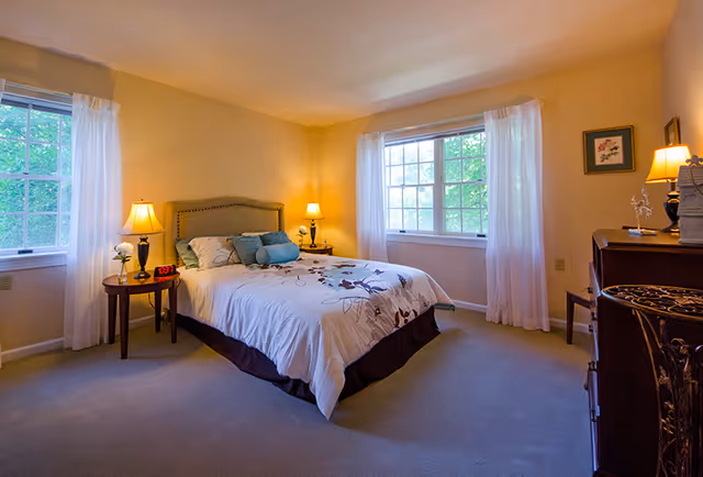 A cozy bedroom with a double bed featuring a floral patterned comforter and multiple pillows. The room has two large windows with white curtains allowing natural light to fill the space. There are two bedside tables with lamps on either side of the bed, a digital clock on one table, and a dresser with a lamp and framed artwork on the wall. The walls are painted a soft yellow and the floor is carpeted.