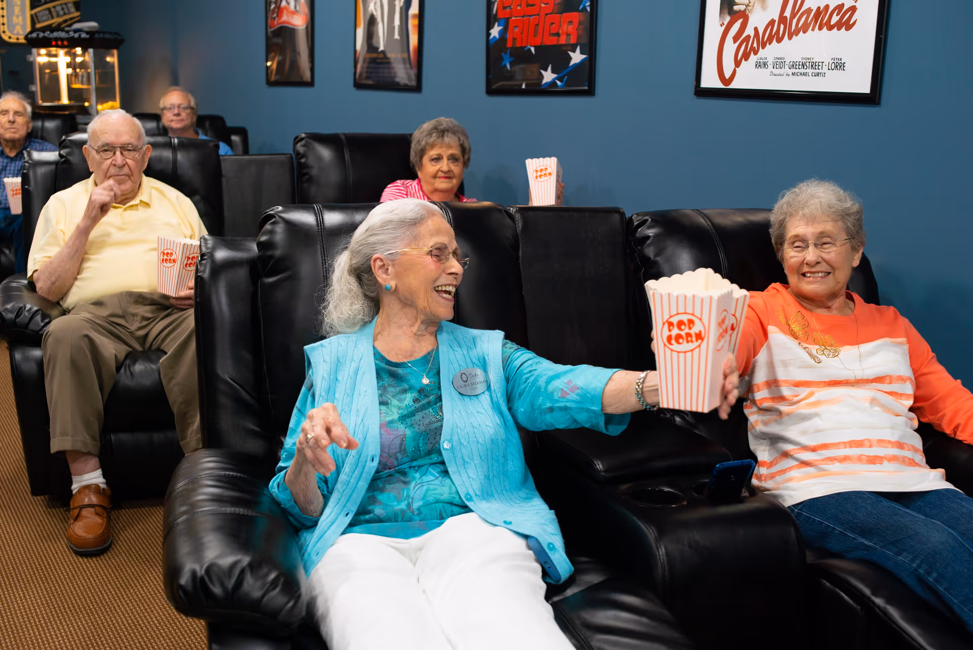A group of elderly people sitting in black leather recliners in a movie theater room. Two women in the front row are smiling and sharing a popcorn container. Other seniors are seated behind them, also holding popcorn. Movie posters are visible on the blue walls.
