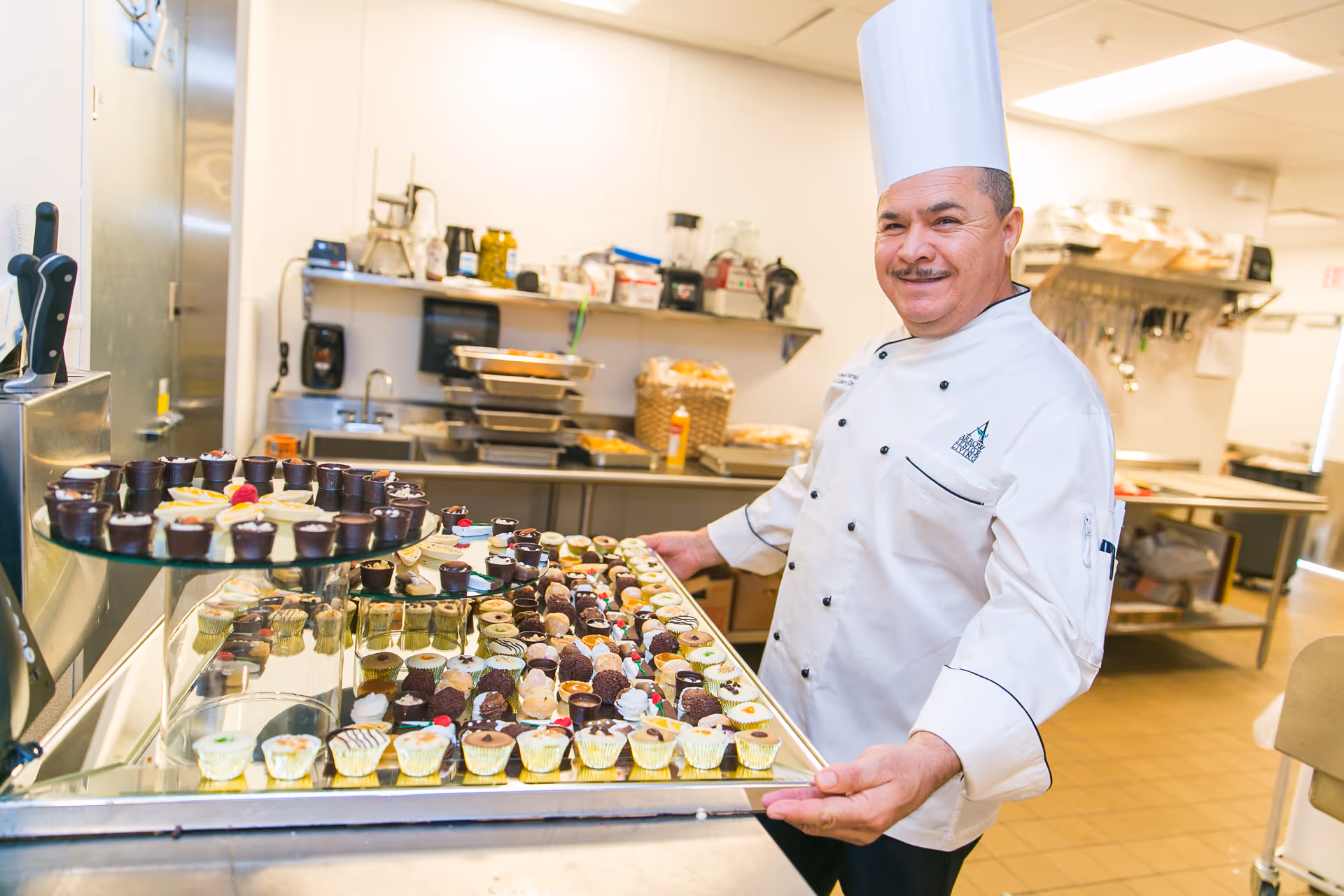 A chef wearing a white chef's coat and tall white chef hat is smiling and holding a large tray filled with assorted mini desserts in a commercial kitchen.