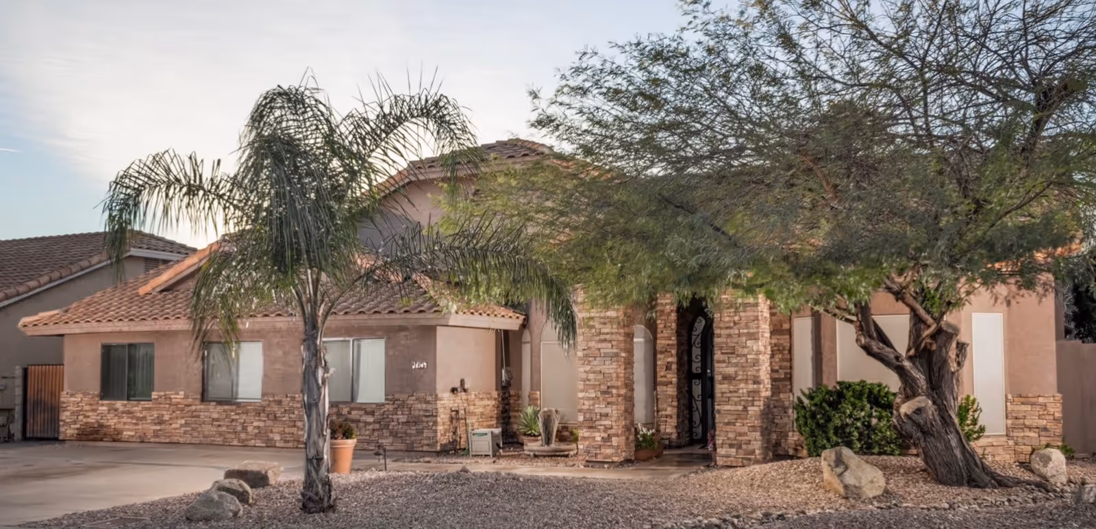 Exterior view of a single-story building with a tiled roof and stone facade accents. There are two trees in front, one palm tree and one larger leafy tree, with rocks and gravel landscaping around the entrance.