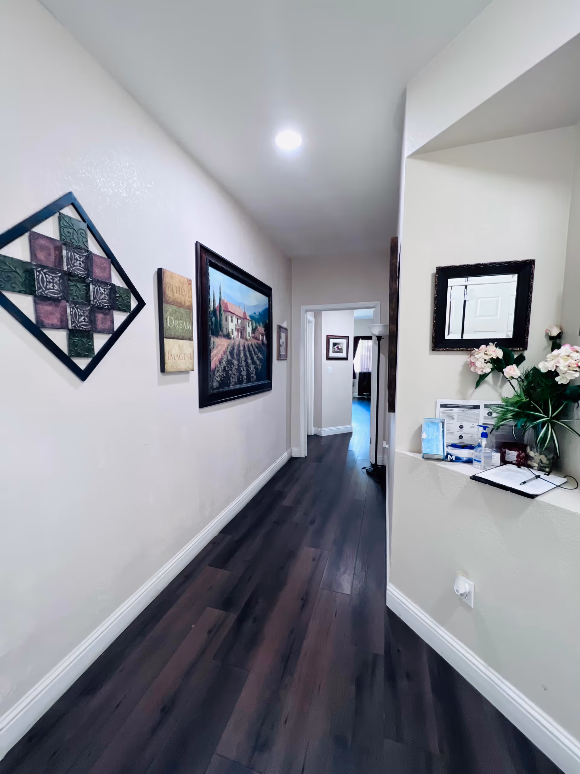 A hallway with dark wooden flooring and light-colored walls decorated with framed artwork and a decorative wall piece. There is a small counter on the right side with flowers, hand sanitizer, and some papers. A mirror hangs above the counter, and the hallway leads to another room with visible wall art.