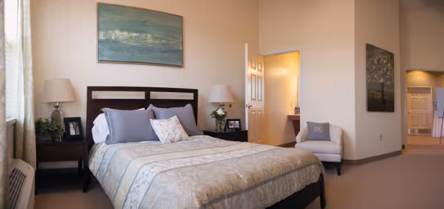 A well-lit bedroom with a large bed featuring a patterned comforter and multiple pillows. There are two nightstands on either side of the bed, each with a lamp and decorative items. A painting hangs above the bed. To the right, an open door leads to a bathroom, and a white armchair is positioned near the corner. The room has beige walls and carpeted floors.