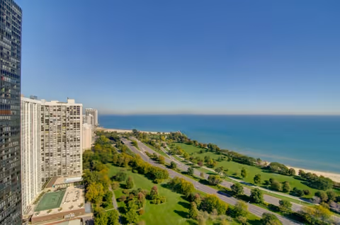A high-angle view of a coastal area showing a large green park with trees and a road running parallel to a sandy beach and blue ocean under a clear sky. Tall buildings are visible on the left side of the image.