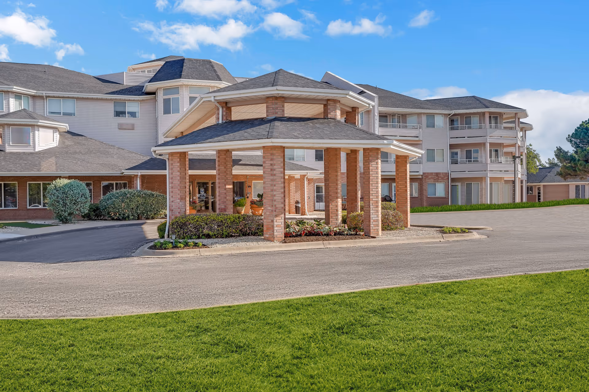 Exterior view of a senior living facility building with a covered entrance supported by brick columns, surrounded by well-maintained landscaping and green grass under a blue sky with some clouds.