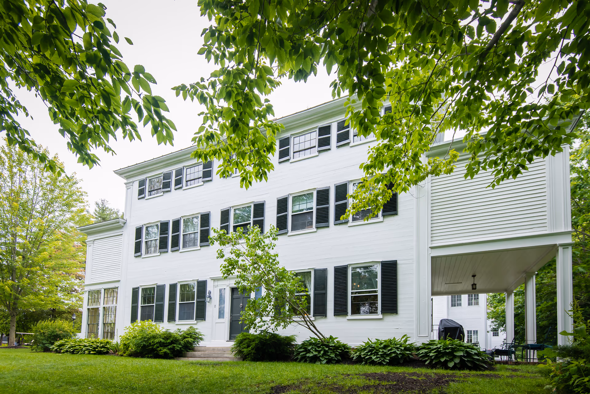A large white multi-story building with black shutters surrounded by green trees and bushes, viewed from the front with a grassy lawn in the foreground.