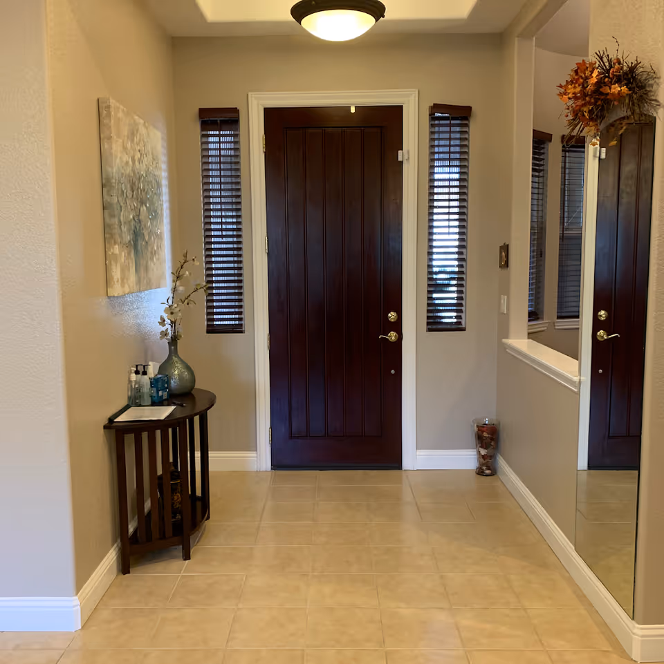 Entryway of a senior living facility with a dark wooden front door flanked by two narrow windows with blinds. To the left, there is a small wooden table with a vase holding white flowers and several bottles of hand sanitizer. A large mirror is mounted on the right wall, reflecting part of the door and window. The floor is tiled, and a decorative glass container with dried flowers is placed in the corner near the mirror. A ceiling light fixture illuminates the area.