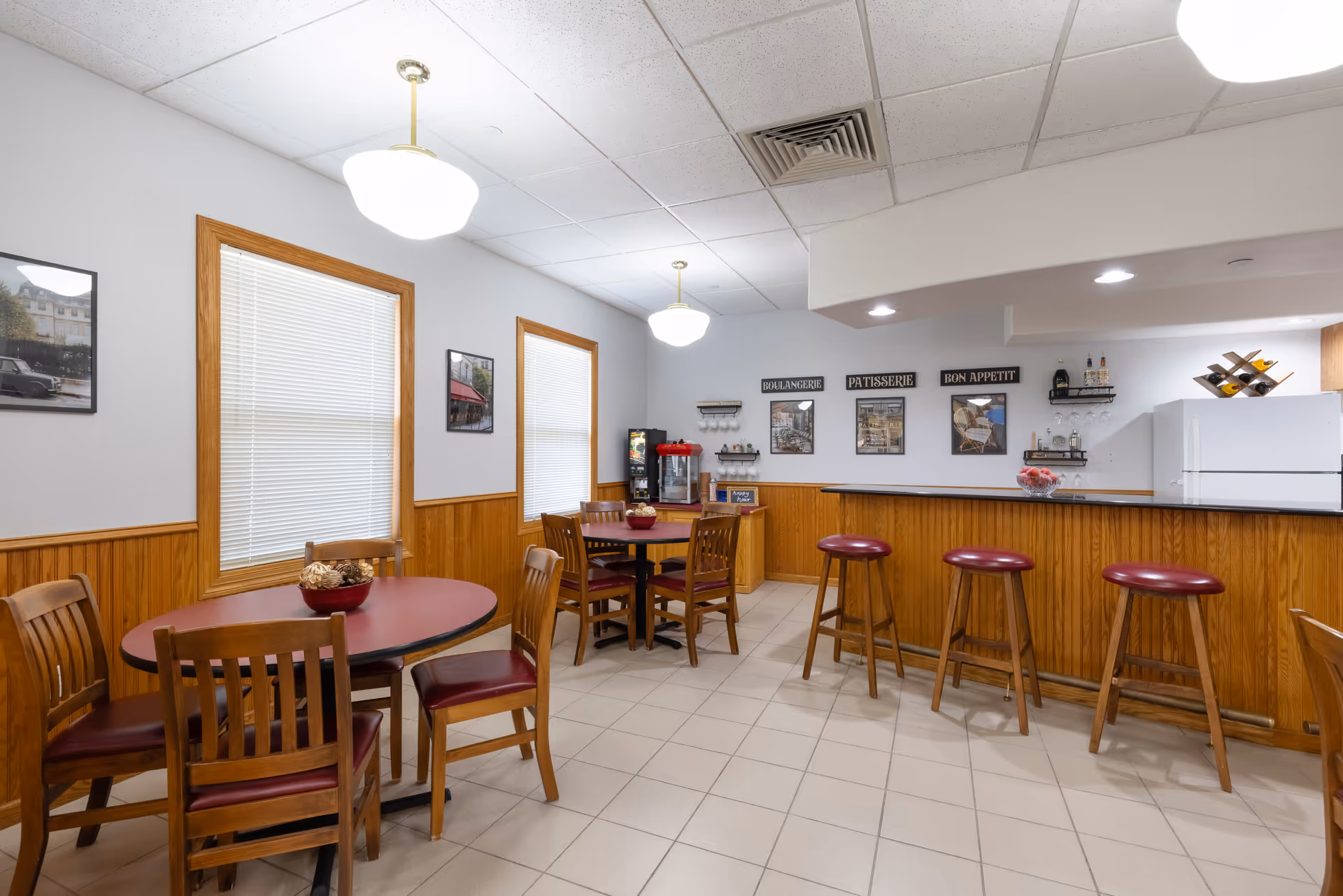 Interior view of a dining area with two round tables surrounded by wooden chairs with red cushions. There is a wooden counter with four red cushioned bar stools. The walls have wood paneling on the lower half and light gray paint above, decorated with framed pictures and small shelves. Two windows with blinds are visible, and the ceiling has white tiles with two hanging light fixtures.