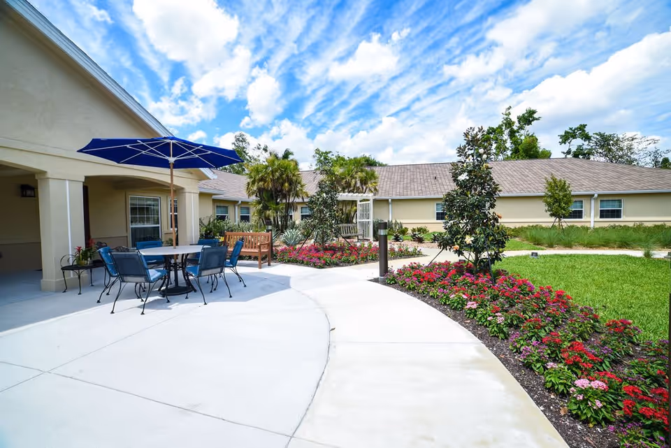 Outdoor patio area at a senior living facility with a round table, blue chairs, and a blue umbrella. There is a wooden bench nearby, surrounded by landscaped flower beds with red and pink flowers, green grass, and small trees. The building is beige with a sloped roof under a partly cloudy blue sky.