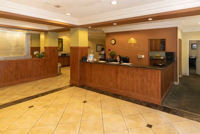 Reception area of The Grove Care and Wellness facility with a wooden front desk, a receptionist seated behind the desk, beige tiled floor, and warm-toned walls with the facility's logo displayed behind the desk.