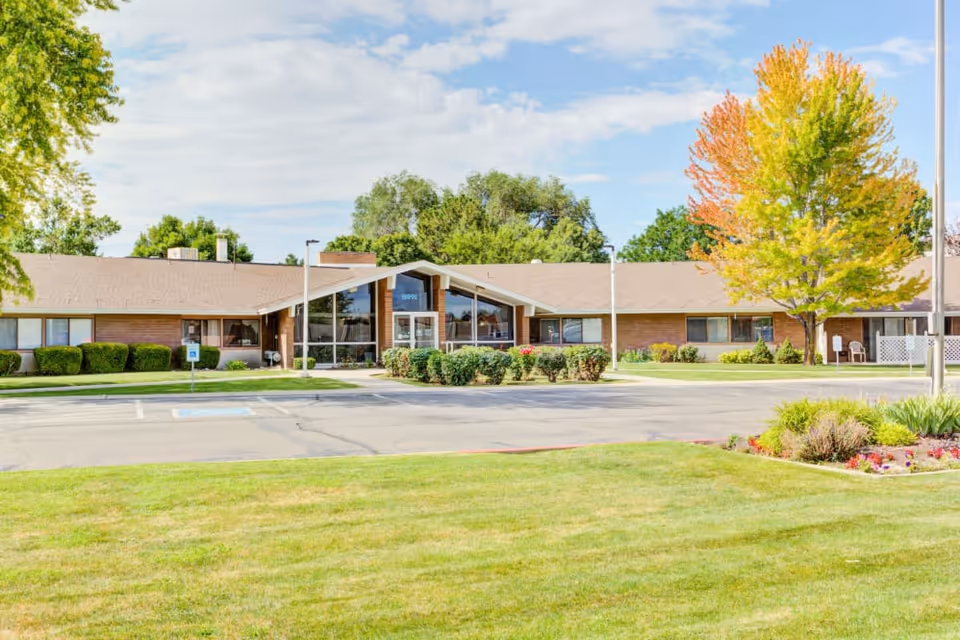 Front view of a single-story brick senior living facility with a landscaped lawn, driveway, and a central glass entrance.