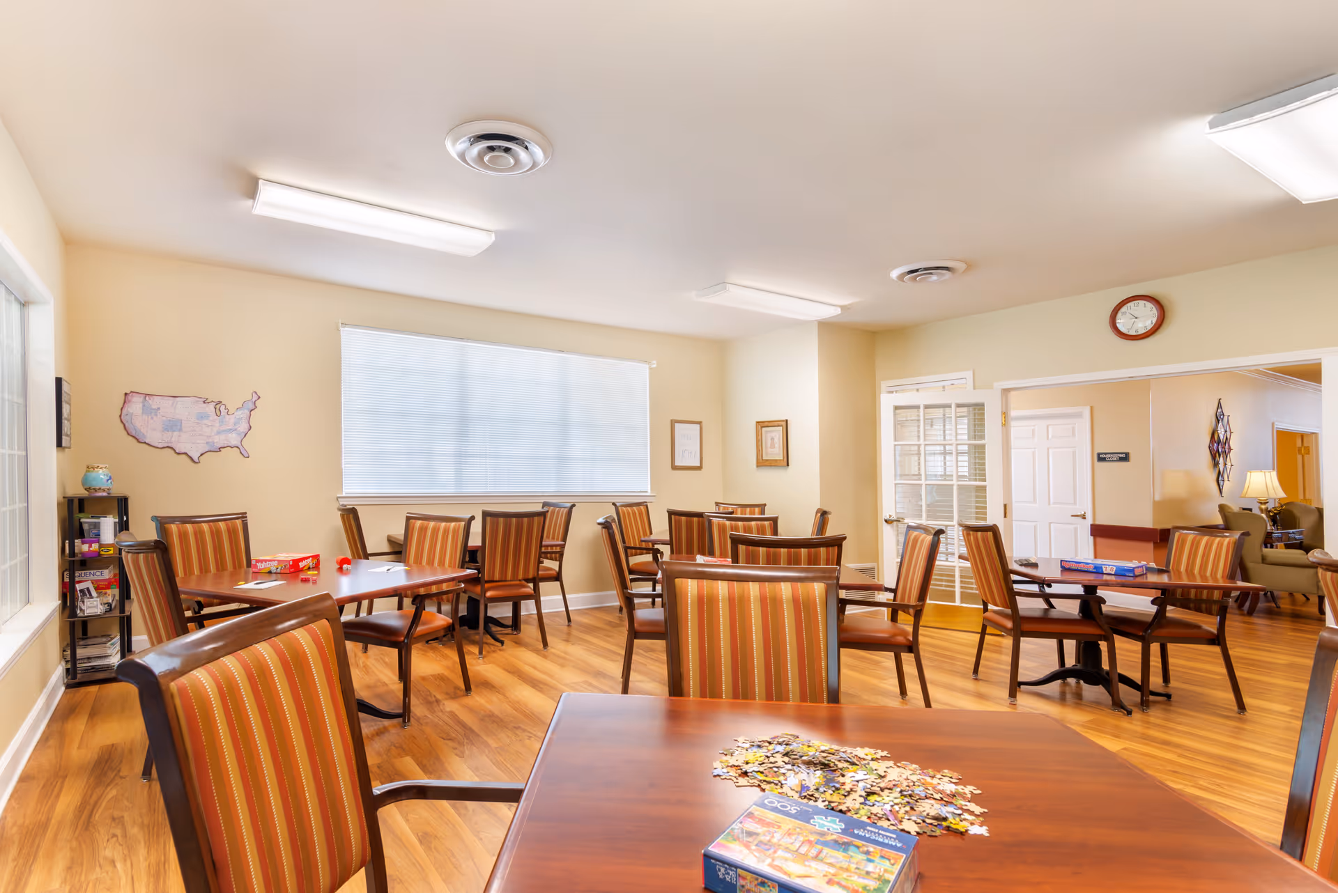 Bright communal activity/dining room with multiple tables and striped chairs, a puzzle and board game on the nearest table, and wood flooring.