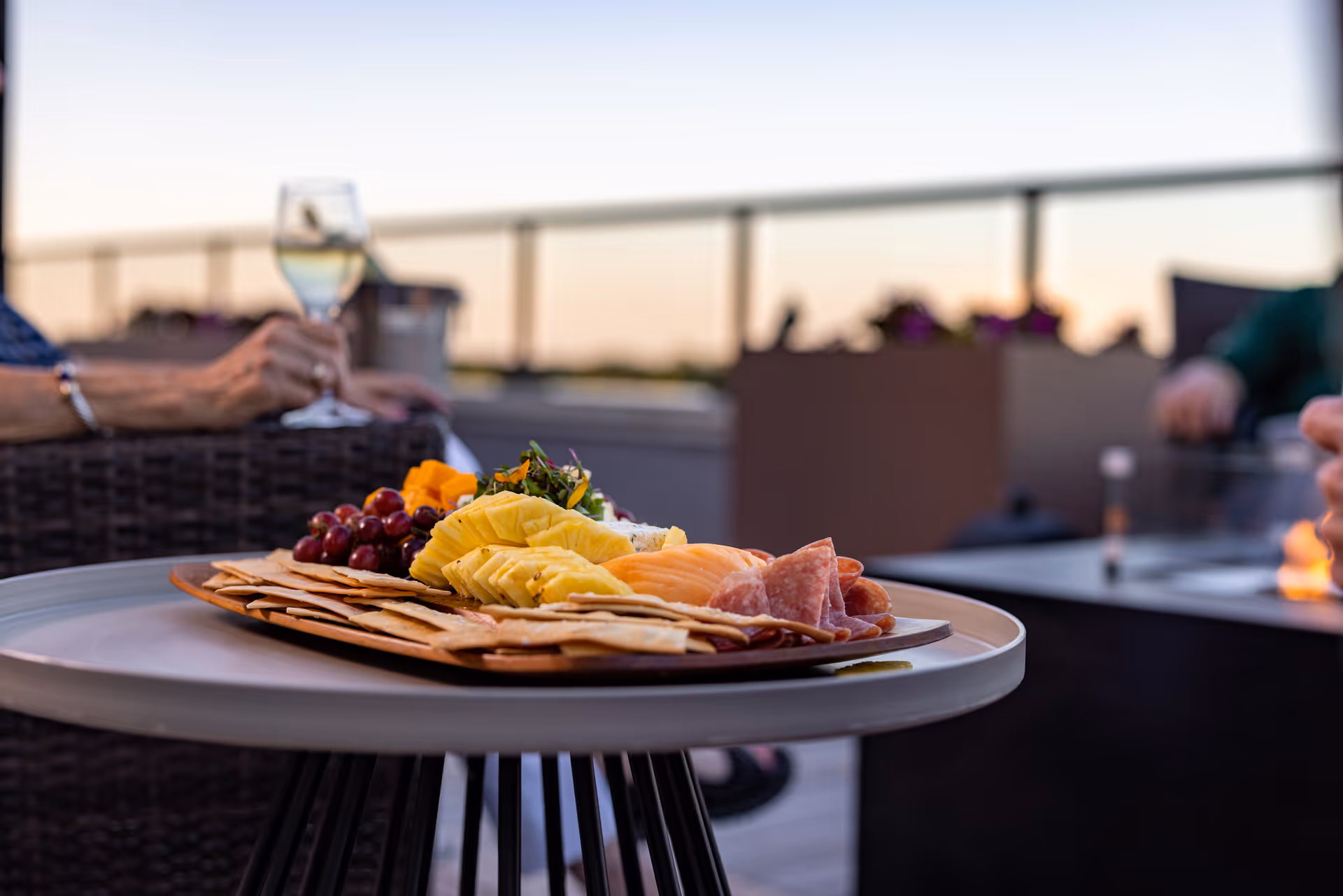 A close-up view of a platter with crackers, sliced pineapple, grapes, cheese, and sliced meats on a round table outdoors. In the background, a person is holding a glass of white wine while sitting on a wicker chair, with a blurred outdoor setting and railing visible.