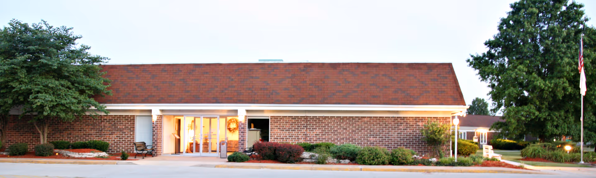 Front exterior view of a single-story brick building with a red shingled roof, large glass entrance doors, and landscaping including trees and shrubs on either side. An American flag is visible on the right side near the building.
