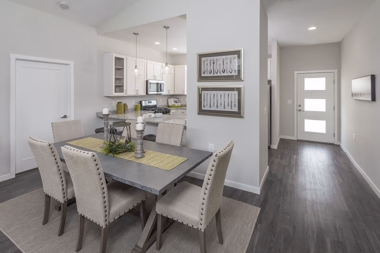 A modern dining area with a rectangular table surrounded by six upholstered chairs with nailhead trim. The table is decorated with a green runner, two candle holders, and a small plant centerpiece. Behind the dining area is a kitchen with white cabinets, granite countertops, a stove, and a microwave. Two pendant lights hang above the kitchen counter. To the right is a hallway leading to a door with three frosted glass panels. The walls are painted light gray and the floor is dark wood.