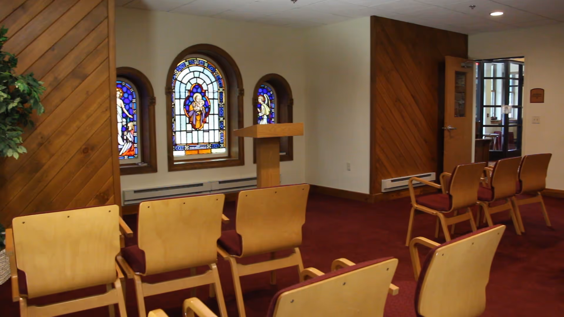 Chapel-like interior with rows of wooden chairs facing a lectern, wood-paneled walls, and three stained-glass windows.