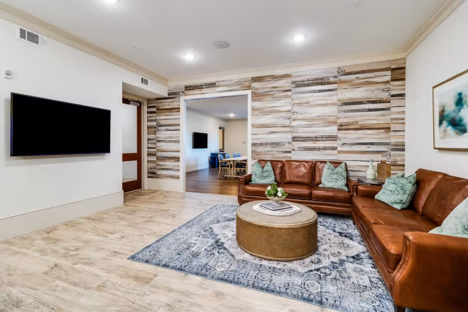 A cozy living room with two brown leather sofas adorned with green pillows, a round wooden coffee table with a small flower arrangement and books on a patterned blue and white rug. The room features a rustic wooden accent wall and a flat-screen TV mounted on a white wall. Through an open doorway, a dining area with a table and chairs is visible.
