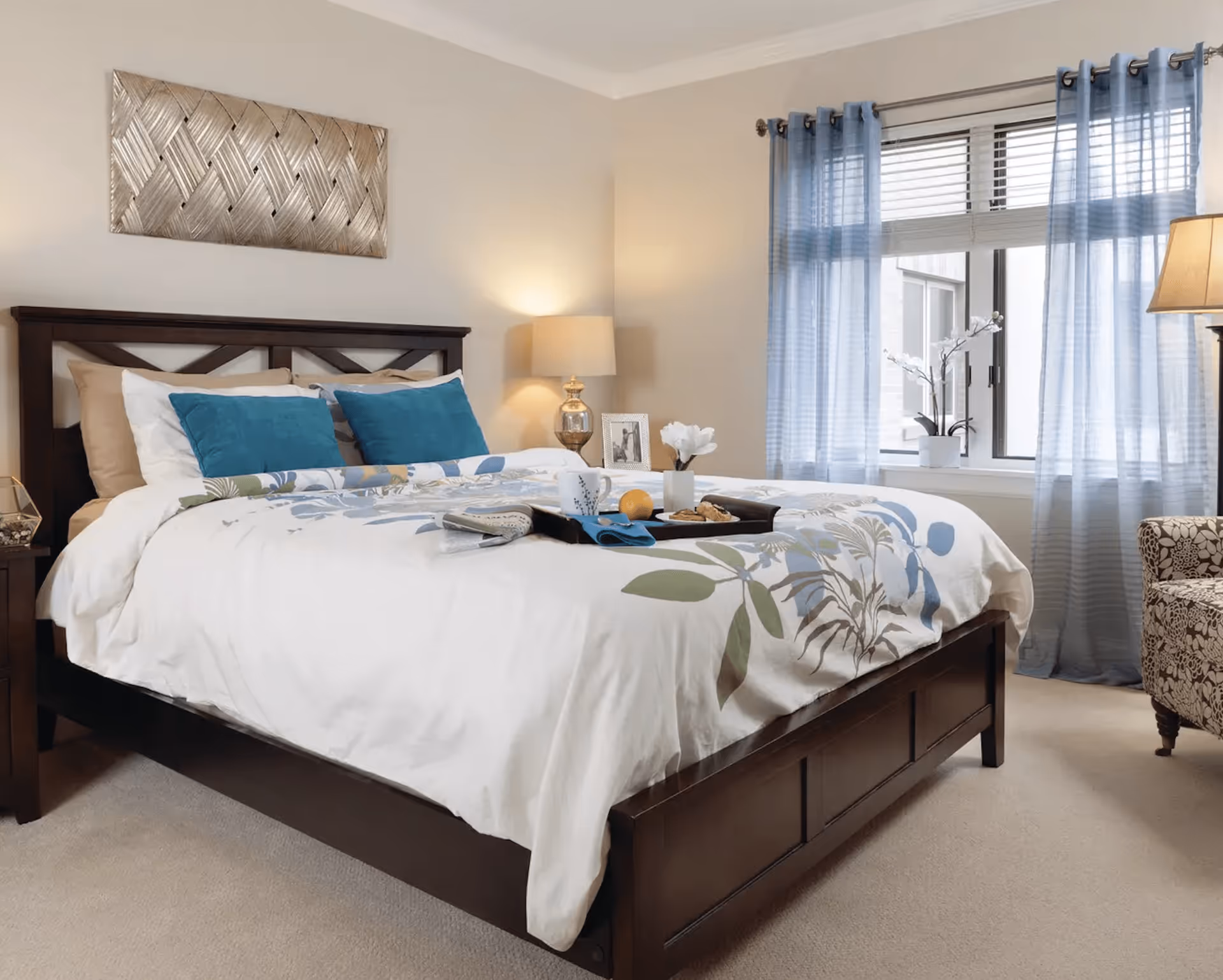 A neatly made bed with a dark wooden frame and floral-patterned white bedding, accented by two blue pillows. A tray with a cup, an orange, and some cookies is placed on the bed. The room has beige walls, a window with blue curtains, a lamp on a bedside table, a framed photo, and a patterned armchair.