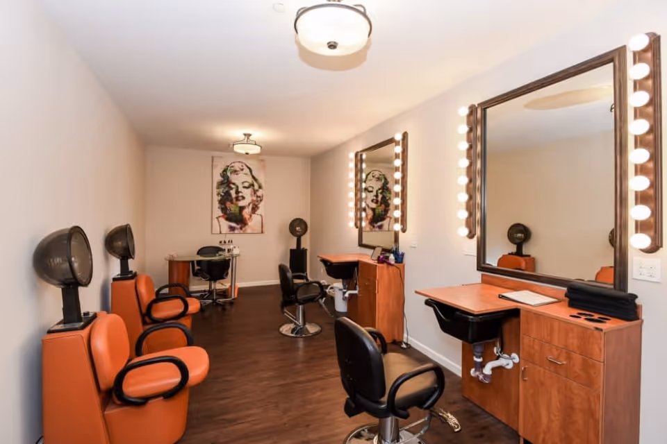 Interior view of a salon room with orange salon chairs, hair dryers, large mirrors with lights around the edges, and a portrait of Marilyn Monroe on the far wall.