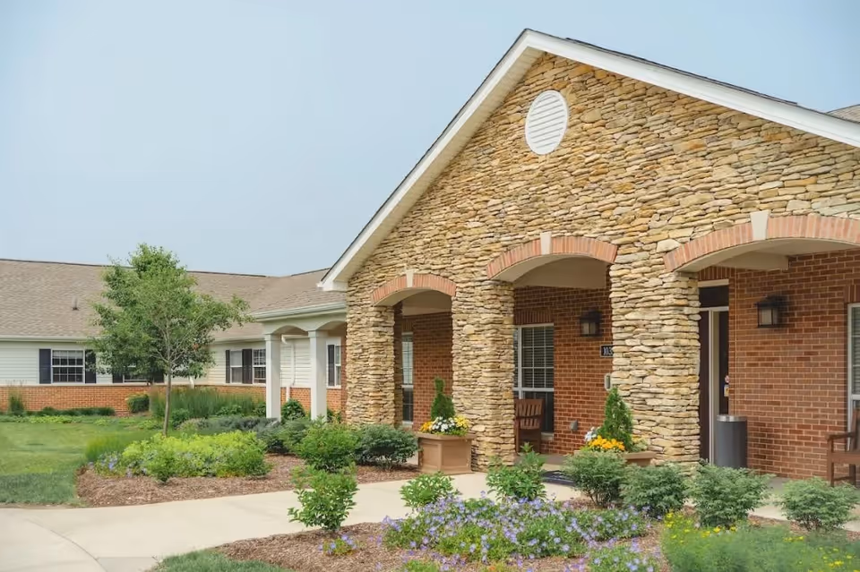 Exterior view of a senior living facility building with a stone and brick facade, arched entryways, and a landscaped garden with shrubs and flowers in front.