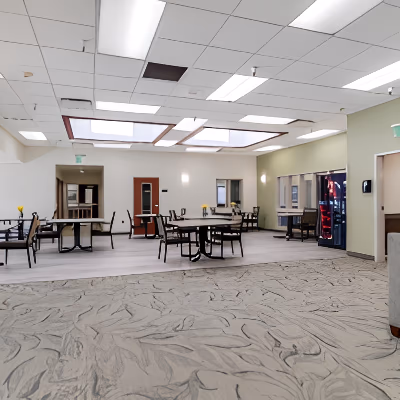 Spacious common dining area with round tables and chairs under skylights and a vending machine along the wall.