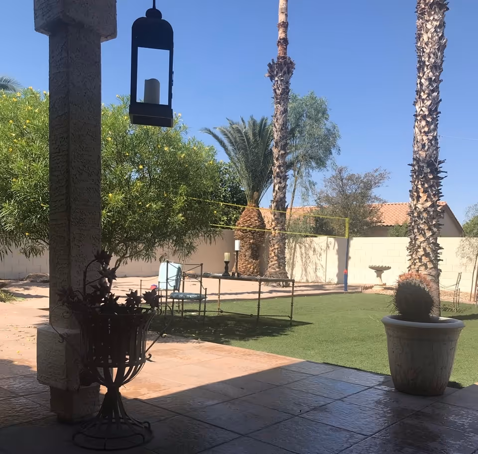 View of an outdoor patio area with tiled flooring, a hanging lantern, and a decorative metal plant stand. Beyond the patio is a grassy area with a volleyball net, palm trees, and other greenery, enclosed by a white brick wall under a clear blue sky.