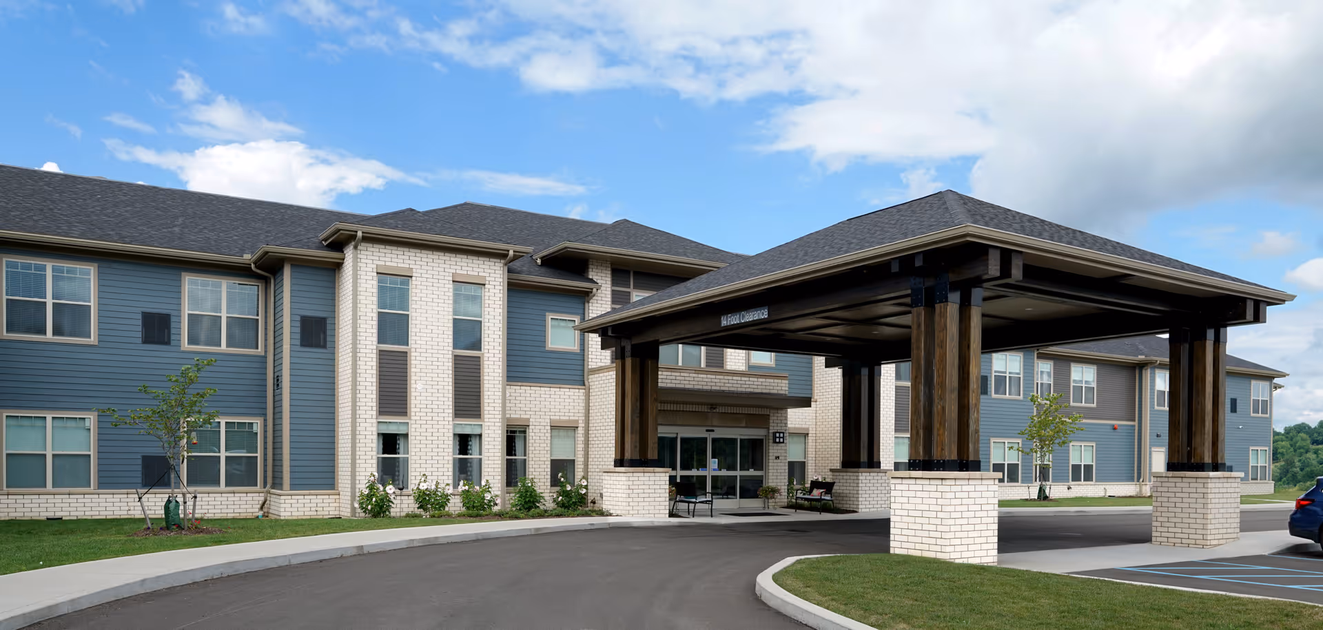 Exterior view of a senior living facility building with a covered entrance driveway. The building has a combination of blue siding and light-colored brick with multiple windows. There is a curved driveway leading to the entrance, some small trees and shrubs planted around the building, and a partly cloudy sky above.