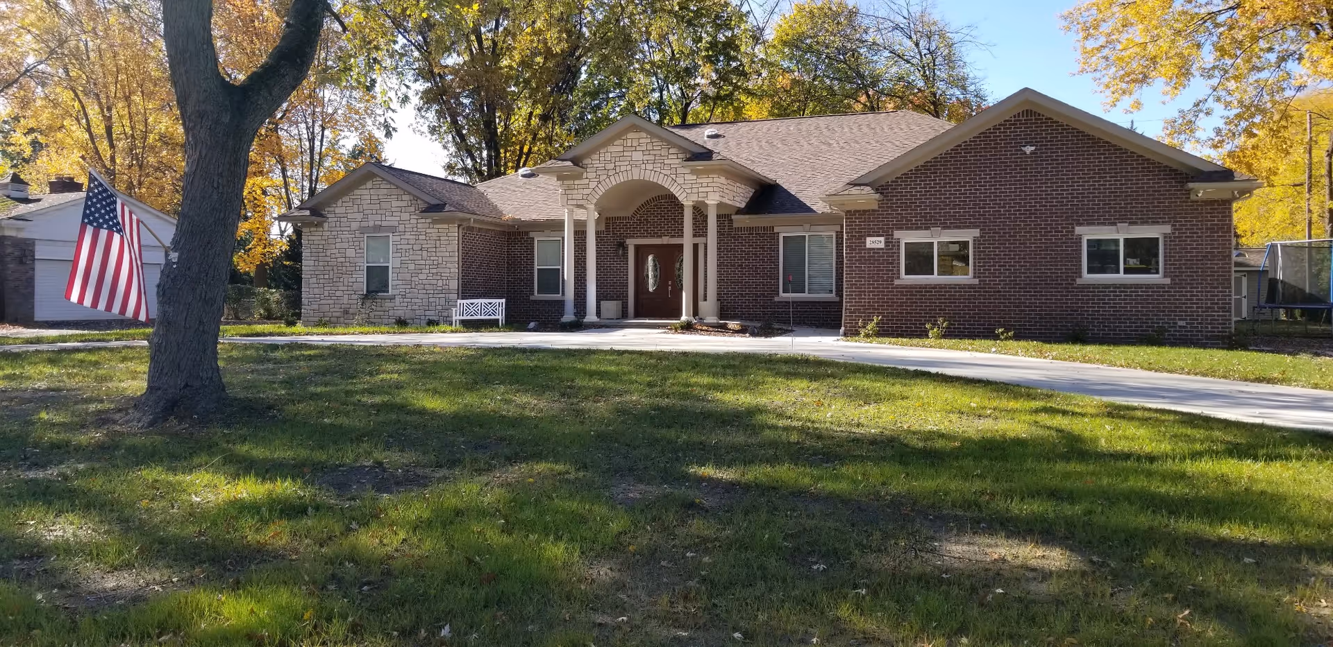 Single-story brick and stone building with a covered front porch, surrounded by green grass and trees with autumn foliage. An American flag is displayed on a pole near a large tree in the front yard. A concrete driveway leads up to the entrance.