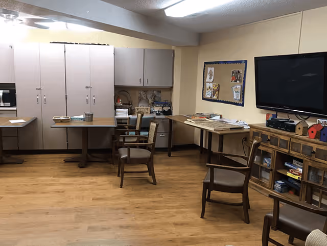 A common room with wooden flooring, several wooden chairs with cushions, tables with various items on them, a large cabinet with closed doors, a bulletin board with pinned pictures, and a flat-screen TV mounted on the wall above a wooden cabinet containing board games and other items.
