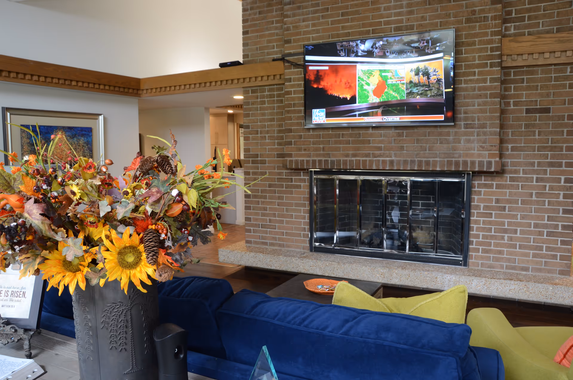 A cozy living room area with a large brick fireplace and a flat-screen TV mounted above it displaying a news channel. In front of the fireplace is a blue couch with yellow and green pillows, and a coffee table with a decorative orange dish. A large vase filled with autumn-themed flowers and pinecones is placed on a side table next to the couch. The room has warm lighting and wooden trim along the ceiling.