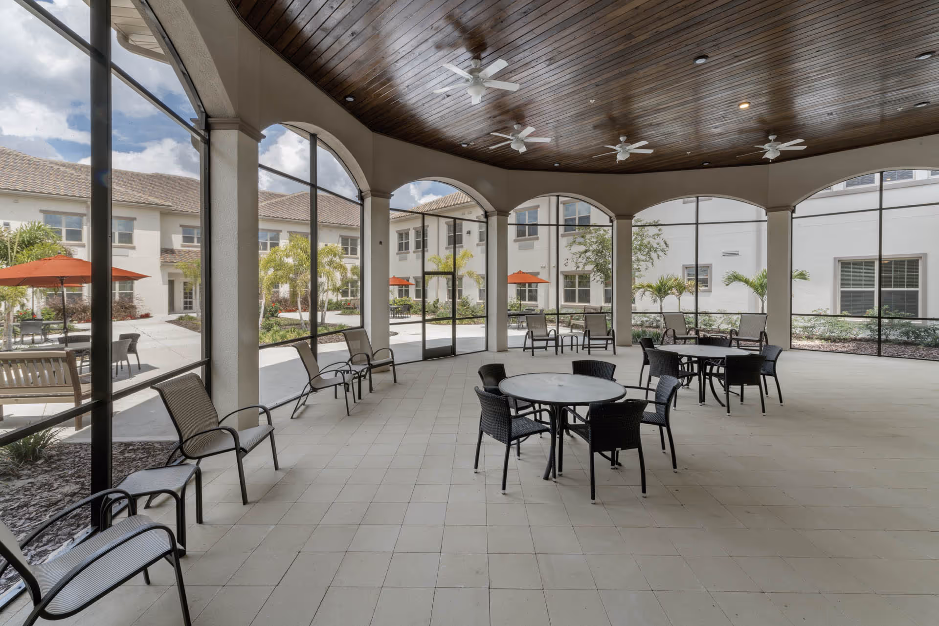 Covered outdoor seating area with round tables and chairs, surrounded by large screened windows and ceiling fans, overlooking a courtyard with palm trees and patio umbrellas.