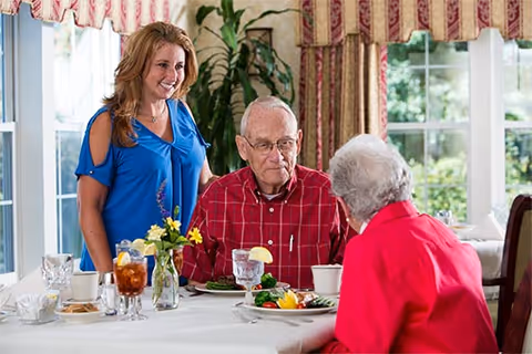 A smiling woman in a blue blouse stands next to an elderly man in a red plaid shirt and an elderly woman in a red jacket, who are seated at a dining table with plates of food and drinks in a well-lit room with large windows and floral curtains.