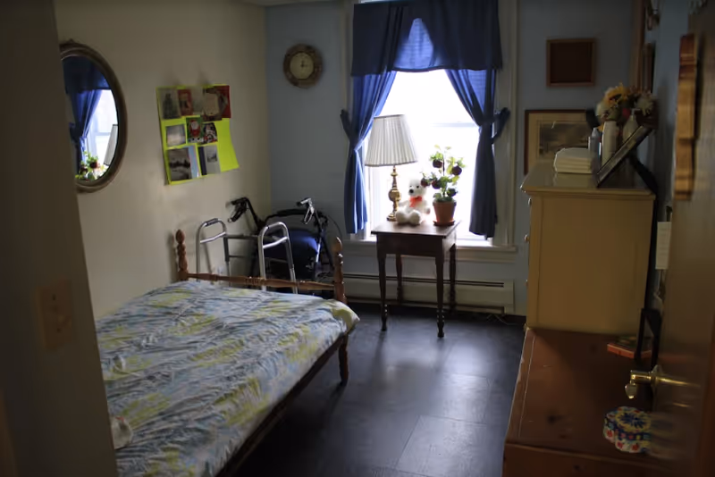 A small bedroom with a single bed covered in a patterned bedspread, a walker and wheelchair beside the bed, a wooden side table with a lamp, potted plant, and a teddy bear in front of a window with blue curtains. There is a dresser with folded towels and framed pictures on top, a round mirror on the wall, and a clock above the window.