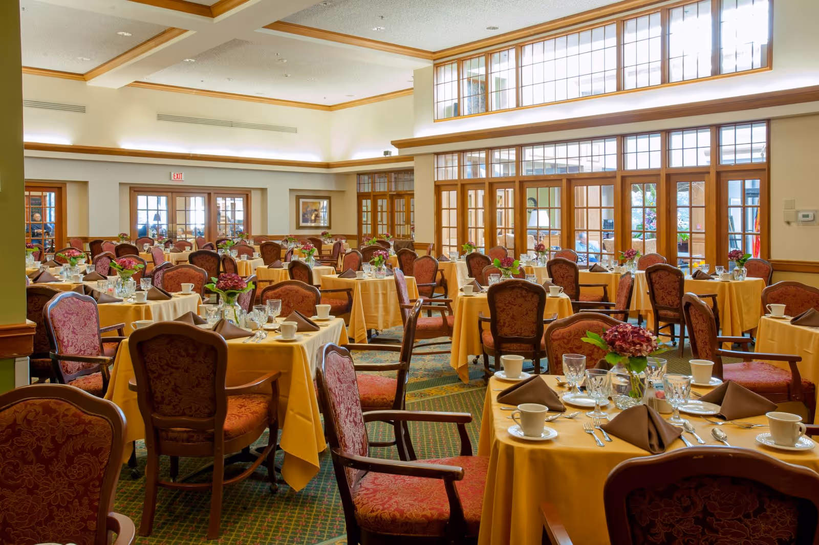 Spacious dining room with round tables draped in yellow tablecloths, set with place settings and floral centerpieces.