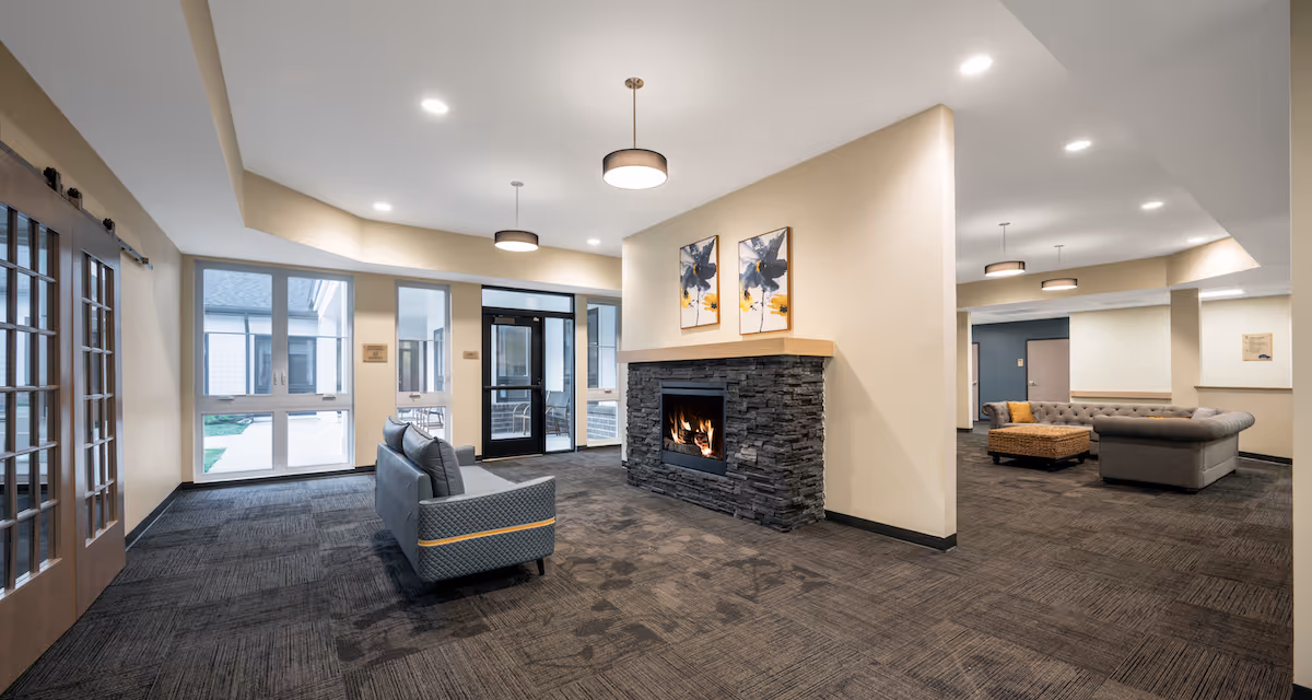 A spacious senior living facility common area with a modern stone fireplace in the center, two gray sofas facing each other, large windows letting in natural light, and pendant ceiling lights. The room has beige walls and dark carpet flooring, with glass doors leading to an outdoor area.
