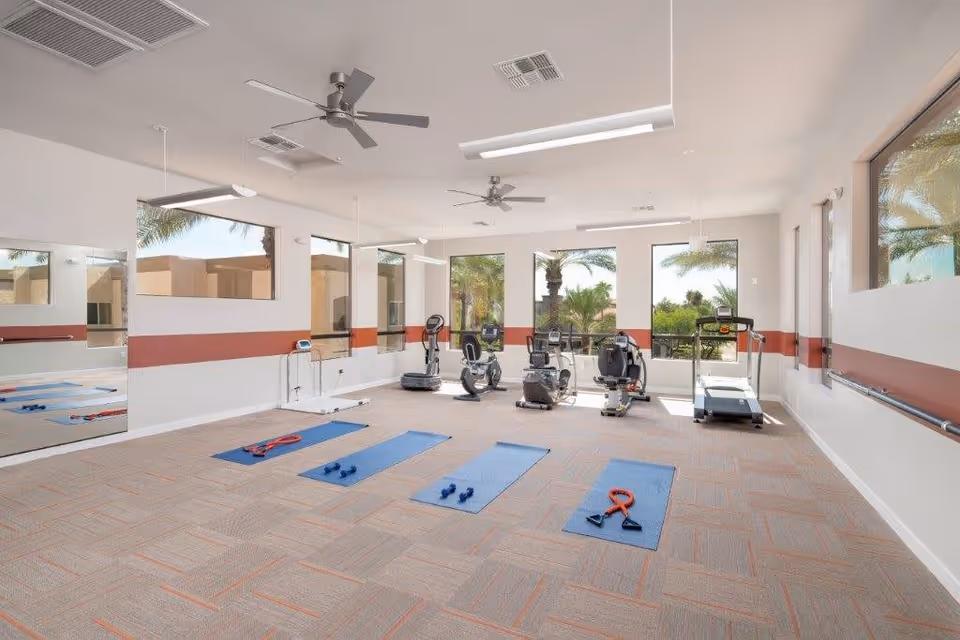 A bright exercise room with large windows showing palm trees outside. The room contains four blue exercise mats on the carpeted floor, each with small dumbbells or resistance bands. There are several pieces of exercise equipment including a treadmill, stationary bikes, and a vibration plate. The walls are white with a red stripe, and there are ceiling fans and modern lighting fixtures.