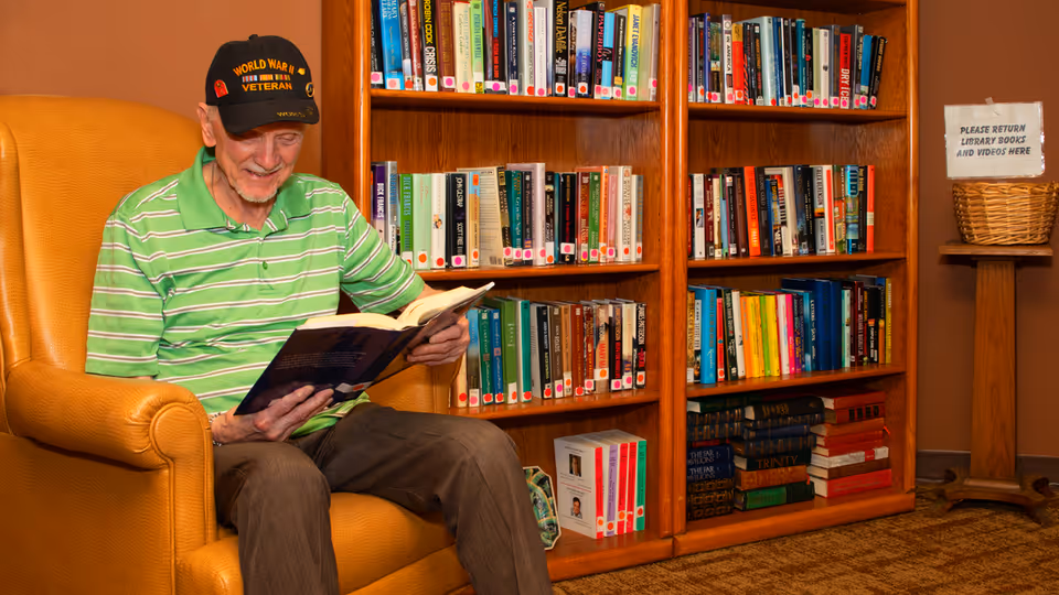 An elderly man wearing a World War II veteran cap and a green striped polo shirt is sitting in a comfortable orange armchair, reading a book. Behind him are wooden bookshelves filled with various books. To the right, there is a small table with a basket and a sign that reads 'PLEASE RETURN LIBRARY BOOKS AND VIDEOS HERE.'
