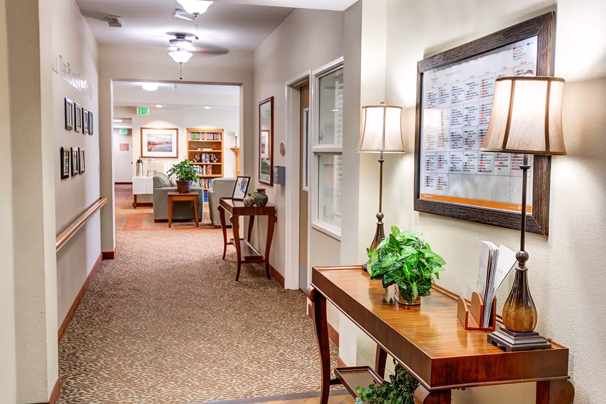 A well-lit interior hallway of a senior living facility with wooden console tables, lamps, plants, and a seating area and bookshelf at the far end.