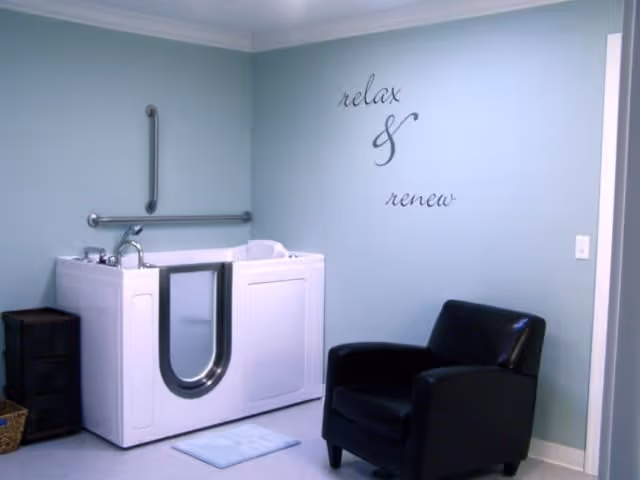 A small room with a white walk-in bathtub equipped with safety grab bars, a black cushioned armchair, and a wall decal that reads 'relax & renew'. The walls are painted light blue and there is a small black storage unit and a basket on the floor.