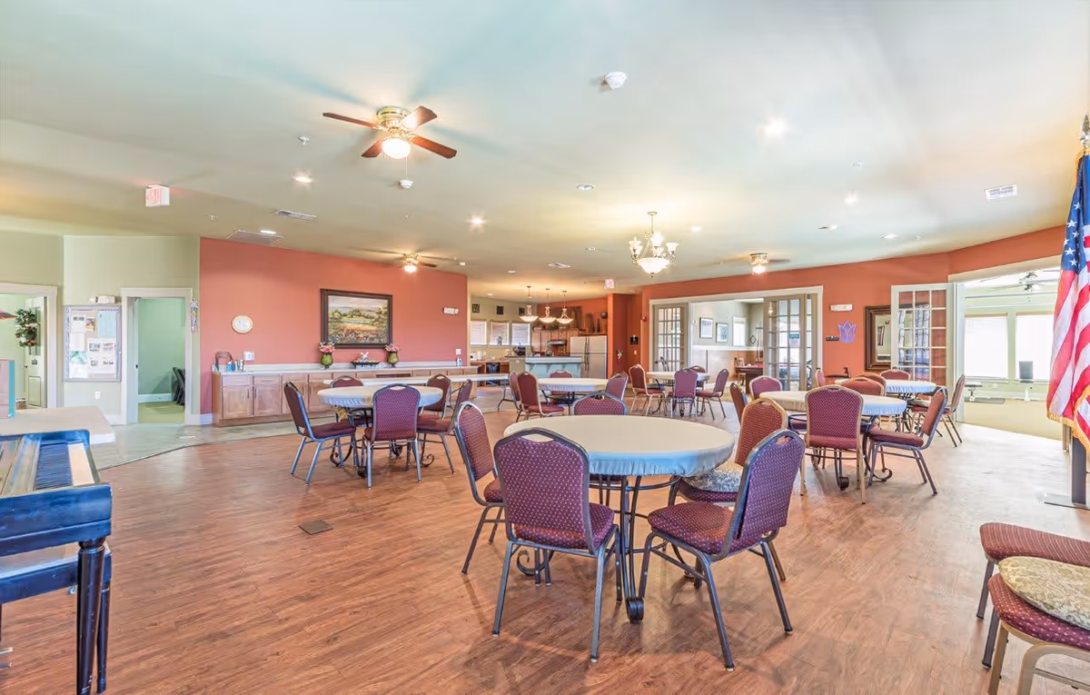 A spacious community room with round tables covered with light blue tablecloths and surrounded by maroon chairs. The room has wooden flooring, ceiling fans, and warm lighting. There is a kitchen area in the background with a refrigerator and cabinets, and an American flag is visible on the right side near the entrance. The walls are painted in a combination of light green and reddish tones, and there are framed pictures and decorations on the walls.