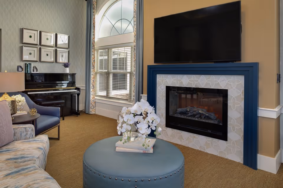 Cozy living room with a wall-mounted TV above a fireplace, a piano by the window, and seating around a round ottoman.